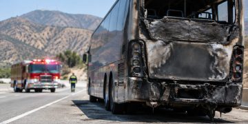 Charred rear of a tour bus on the shoulder of a freeway in Burbank in morning daylight, with a fire engine blurred in the background.