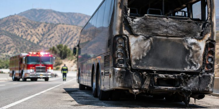 Charred rear of a tour bus on the shoulder of a freeway in Burbank in morning daylight, with a fire engine blurred in the background.