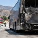 Charred rear of a tour bus on the shoulder of a freeway in Burbank in morning daylight, with a fire engine blurred in the background.