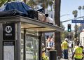 Close-up of a Los Angeles bus shelter with personal belongings on its roof, with city workers and the Brentwood street scene blurred in the background.