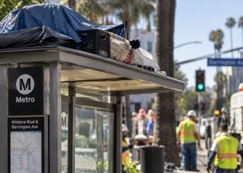 Close-up of a Los Angeles bus shelter with personal belongings on its roof, with city workers and the Brentwood street scene blurred in the background.
