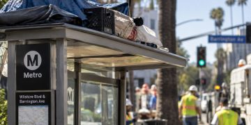 Close-up of a Los Angeles bus shelter with personal belongings on its roof, with city workers and the Brentwood street scene blurred in the background.