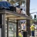 Close-up of a Los Angeles bus shelter with personal belongings on its roof, with city workers and the Brentwood street scene blurred in the background.