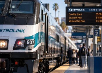 Close-up of a Metrolink train at a Southern California station, with a digital fare display and waiting passengers softly blurred in the background.