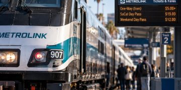 Close-up of a Metrolink train at a Southern California station, with a digital fare display and waiting passengers softly blurred in the background.