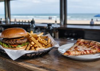Close-up of a burger with fries in a paper-lined basket and a pizza slice on a wooden bar counter, background softly blurred.
