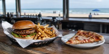 Close-up of a burger with fries in a paper-lined basket and a pizza slice on a wooden bar counter, background softly blurred.