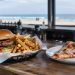 Close-up of a burger with fries in a paper-lined basket and a pizza slice on a wooden bar counter, background softly blurred.