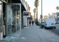 Close-up of a cafe glass door with shattered glass on the sidewalk in early-morning light, with a blurred Los Angeles street behind it.