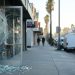 Close-up of a cafe glass door with shattered glass on the sidewalk in early-morning light, with a blurred Los Angeles street behind it.