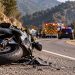 Close-up of a damaged motorcycle on Angeles Crest Highway, with mountain curves, emergency vehicles and rescue activity softly blurred in the background.