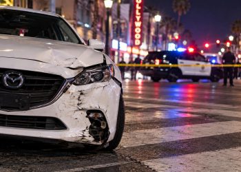 Close-up of a damaged white Mazda stopped on Hollywood Boulevard near a marked pedestrian crosswalk, with police activity softly blurred in the background.