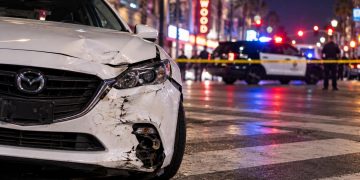 Close-up of a damaged white Mazda stopped on Hollywood Boulevard near a marked pedestrian crosswalk, with police activity softly blurred in the background.