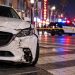 Close-up of a damaged white Mazda stopped on Hollywood Boulevard near a marked pedestrian crosswalk, with police activity softly blurred in the background.