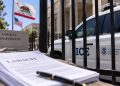 Close-up of a federal immigration enforcement vehicle behind a courthouse fence, with blurred California flags and legal documents in the foreground.