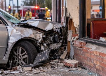 Close-up of a heavily damaged vehicle that crashed into the exterior of a neighborhood restaurant, with emergency responders blurred in the background.