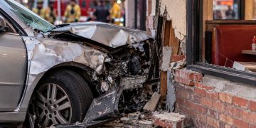 Close-up of a heavily damaged vehicle that crashed into the exterior of a neighborhood restaurant, with emergency responders blurred in the background.