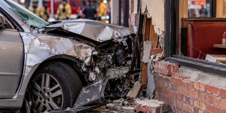 Close-up of a heavily damaged vehicle that crashed into the exterior of a neighborhood restaurant, with emergency responders blurred in the background.