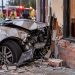 Close-up of a heavily damaged vehicle that crashed into the exterior of a neighborhood restaurant, with emergency responders blurred in the background.
