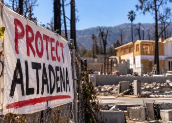 Close-up of a protest banner outside a wildfire-damaged Altadena property, with a partially rebuilt home and burned trees blurred in the background.