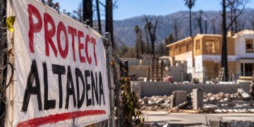Close-up of a protest banner outside a wildfire-damaged Altadena property, with a partially rebuilt home and burned trees blurred in the background.