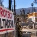 Close-up of a protest banner outside a wildfire-damaged Altadena property, with a partially rebuilt home and burned trees blurred in the background.