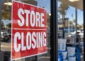 Close-up of a red “STORE CLOSING” sign taped to a retail storefront window, with pool supply products and the parking lot softly blurred in the background.
