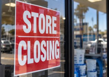 Close-up of a red “STORE CLOSING” sign taped to a retail storefront window, with pool supply products and the parking lot softly blurred in the background.