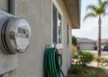 Close-up of a residential electric meter on a stucco wall of a single-story Los Angeles home, with a garden hose and palm tree softly blurred behind.