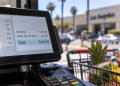 Close-up of a retail checkout terminal displaying a sales total, with a shopping cart and blurred Los Angeles County storefront in the background.