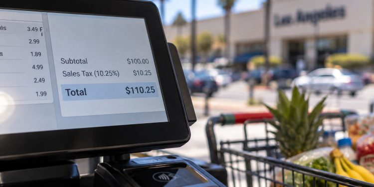 Close-up of a retail checkout terminal displaying a sales total, with a shopping cart and blurred Los Angeles County storefront in the background.