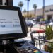 Close-up of a retail checkout terminal displaying a sales total, with a shopping cart and blurred Los Angeles County storefront in the background.