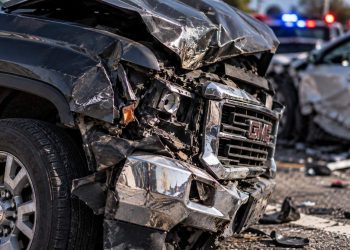 Close-up of a severely damaged GMC pickup truck with a crushed front end and shattered headlight, with blurred emergency vehicles and another wrecked car in the background.