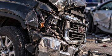 Close-up of a severely damaged GMC pickup truck with a crushed front end and shattered headlight, with blurred emergency vehicles and another wrecked car in the background.