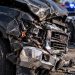 Close-up of a severely damaged GMC pickup truck with a crushed front end and shattered headlight, with blurred emergency vehicles and another wrecked car in the background.