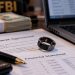 Close-up of a smart ring resting on investment and financial documents beside a laptop, cash and an investigative backdrop, illustrating a federal wearable-technology fraud case.