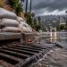 Close-up of a storm drain and sandbags along a Los Angeles street, with dark rain clouds and a hillside neighborhood softly blurred in the background.