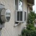 Close-up of an electric meter on the stucco wall of a Los Angeles home, with a window air conditioner softly blurred behind it in daylight.