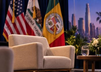 Close-up of an empty beige armchair on a Los Angeles civic forum stage, with U.S., California and Los Angeles flags blurred in the background.