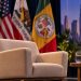 Close-up of an empty beige armchair on a Los Angeles civic forum stage, with U.S., California and Los Angeles flags blurred in the background.