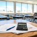 Close-up of budget documents with illegible charts and a calculator on a desk, with a blurred school classroom behind, illustrating the LAUSD budget crisis.