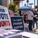 Close-up of campaign signs and voting materials on a Los Angeles sidewalk, with blurred Latino community outreach activity in the background.