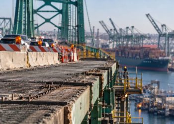 Close-up of the Vincent Thomas Bridge roadway and structural deck, with construction barriers and heavy port traffic blurred in the background.