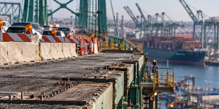 Close-up of the Vincent Thomas Bridge roadway and structural deck, with construction barriers and heavy port traffic blurred in the background.