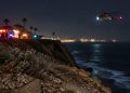 Coastal bluff edge at Point Fermin in San Pedro at night, with loose rocks and scrub in the foreground and a rescue helicopter blurred in the background.