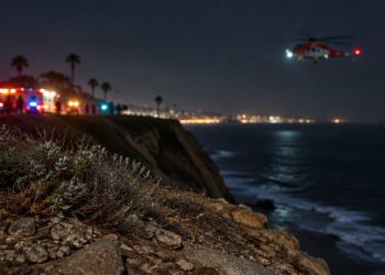 Coastal bluff edge at Point Fermin in San Pedro at night, with loose rocks and scrub in the foreground and a rescue helicopter blurred in the background.