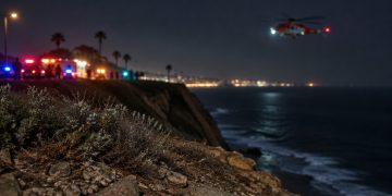 Coastal bluff edge at Point Fermin in San Pedro at night, with loose rocks and scrub in the foreground and a rescue helicopter blurred in the background.