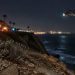 Coastal bluff edge at Point Fermin in San Pedro at night, with loose rocks and scrub in the foreground and a rescue helicopter blurred in the background.
