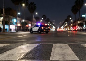 Crosswalk markings glow white in the immediate foreground of a nighttime Broadway street in Florence, with a patrol SUV blurred in the middle distance.