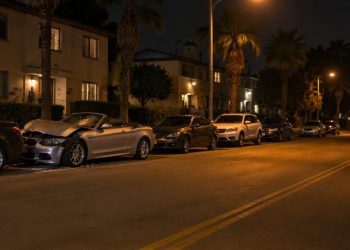 Damaged silver convertible pressed against a row of parked cars on a residential street of apartment buildings and palm trees in University Park at night.
